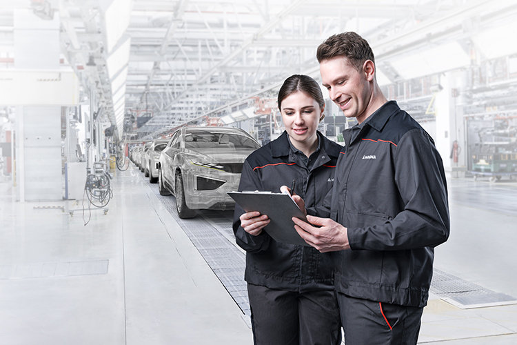 Two people standing holding a folder as finished cars are rolling off the assembly line.