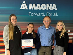 Group of standing together in front of Magna logo and tagline with person in the middle holding a certificate