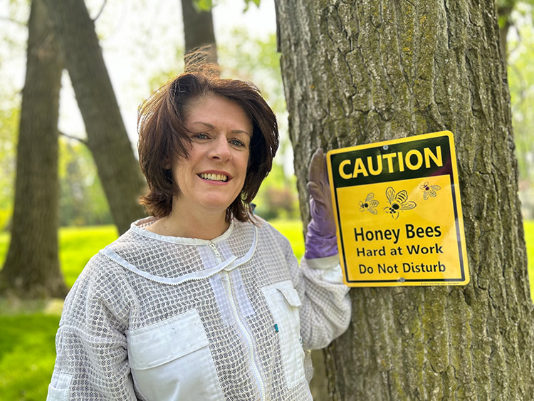 Rhiannon LaForest standing beside a Caution - Honey Bees sign on a tree