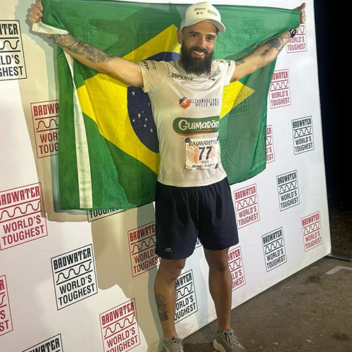 Tiarles Dos Santos standing in front of a step and repeat and holding up a Brazilian flag